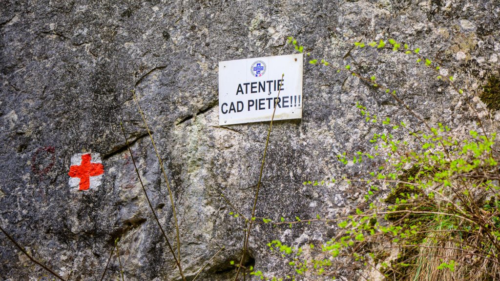 “Caution, falling rocks” sign and red cross trail marking on a stone wall in Turda Gorge